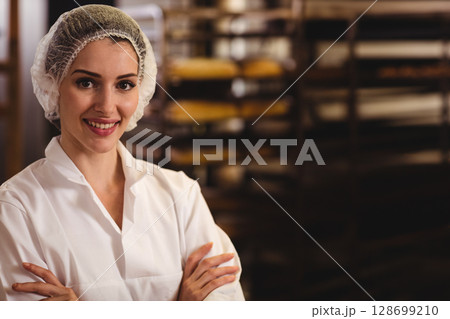 Smiling woman baker standing inside bakery with hairnet and racks of trays, copy space 128699210