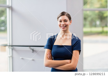 Smiling woman in twenties standing in lobby near large glass window pane and frame, arms crossed 128699211
