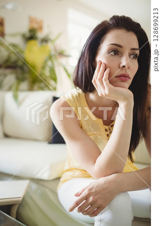 Leaning woman resting chin on hand in modern living room, with laptop and potted plant 128699213