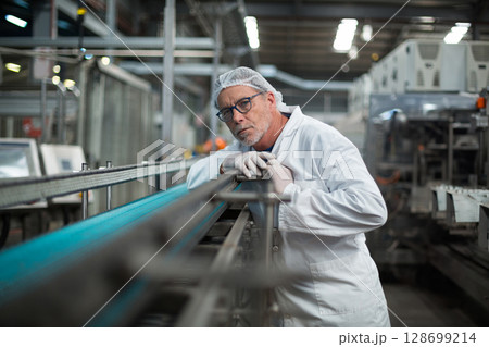 Senior man wearing lab coat and hairnet leaning on conveyor belt railing in factory, with machinery 128699214