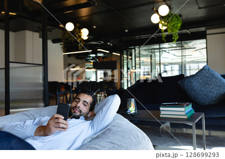 Lounging man reclining on gray beanbag chair in coworking lounge, holding smartphone Lounging man reclining on gray beanbag chair in coworking lounge, holding smartphone 128699293