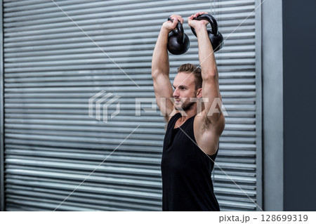 Lifting athletic man pressing two kettlebells overhead before garage door, with industrial theme 128699319