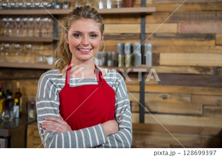 Smiling woman wearing red apron standing in café, with wooden shelves, glassware and spice jars Smiling woman wearing red apron standing in café, with wooden shelves, glassware and spice jars 128699397