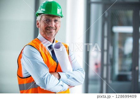 Inspecting man wearing hard hat and safety vest inside lobby, holding blueprints, copy space 128699409