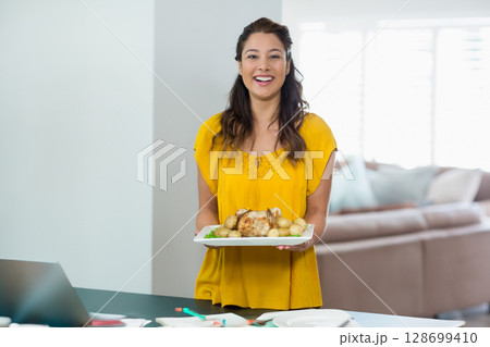 Smiling woman presenting roast chicken platter on kitchen counter, with laptop and papers Smiling woman presenting roast chicken platter on kitchen counter, with laptop and papers 128699410