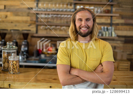 Smiling male barista standing behind coffee shop counter, with glass jars and espresso machine 128699413