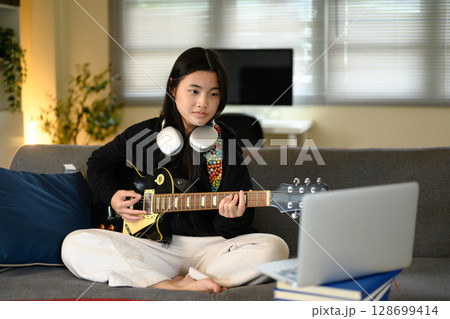 Teenage girl playing electric guitar at home while learning online 128699414