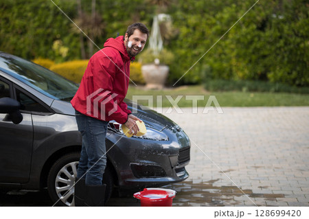 Scrubbing man cleaning hood of hatchback on driveway, with yellow sponge and red bucket, copy space Scrubbing man cleaning hood of hatchback on driveway, with yellow sponge and red bucket, copy space 128699420