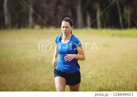 Running athletic woman crossing open grassy field at forest edge, with running shoes 128699421