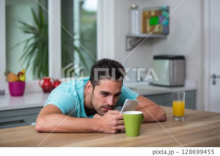 Leaning man scrolling smartphone on kitchen island, with coffee mug and glass of orange juice Leaning man scrolling smartphone on kitchen island, with coffee mug and glass of orange juice 128699455