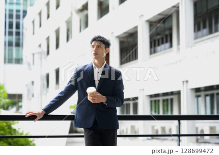 Confident office worker taking a break with coffee in hand against a modern city backdrop Confident office worker taking a break with coffee in hand against a modern city backdrop 128699472