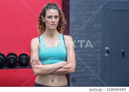 Standing woman crossing arms at gym weight area, with loaded dumbbell rack against red wall Standing woman crossing arms at gym weight area, with loaded dumbbell rack against red wall 128699499