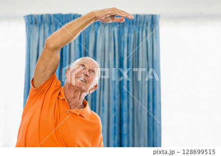 Stretching senior man wearing orange polo shirt lifting arm at home by blue curtains, copy space 128699515