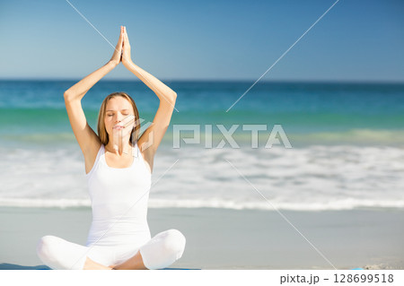 Woman practicing yoga in prayer pose on sandy beach facing ocean, with blue yoga mat 128699518