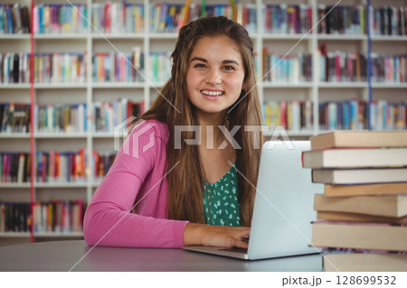 Studying teenage girl sitting at library table, using laptop with book stack and bookshelves Studying teenage girl sitting at library table, using laptop with book stack and bookshelves 128699532