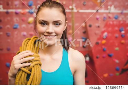 Smiling woman climber holding coiled yellow rope indoors at climbing wall, with colorful handholds 128699554