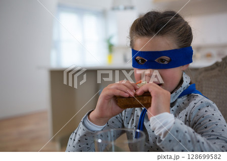 Biting girl child wearing blue mask and cape sitting at kitchen table, with sandwich, copy space Biting girl child wearing blue mask and cape sitting at kitchen table, with sandwich, copy space 128699582