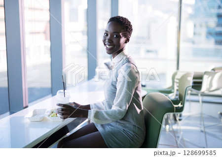 Smiling African American woman sipping from plastic cup at cafeteria counter, with salad container 128699585