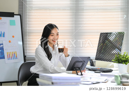 Businesswoman drinking coffee while analyzing financial data on digital tablet in modern office 128699619