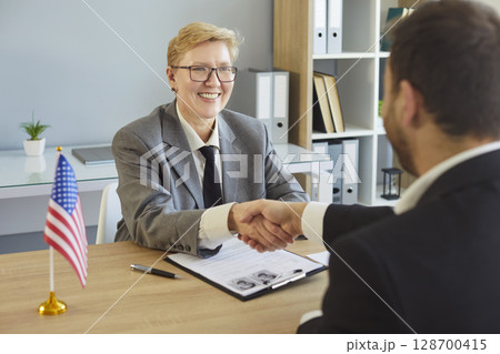 Shaking hands across desk, people sitting greeting in office near American flag, woman smiling Shaking hands across desk, people sitting greeting in office near American flag, woman smiling 128700415