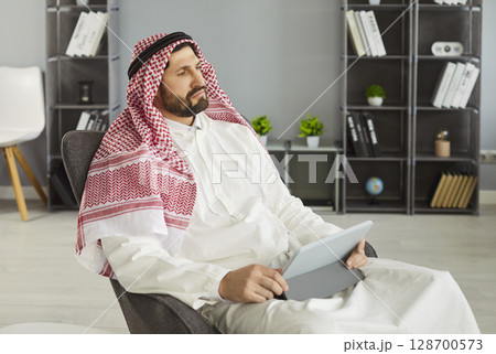 Thoughtful Arabian man posing in armchair with digital tablet, surfing internet or working online, 128700573