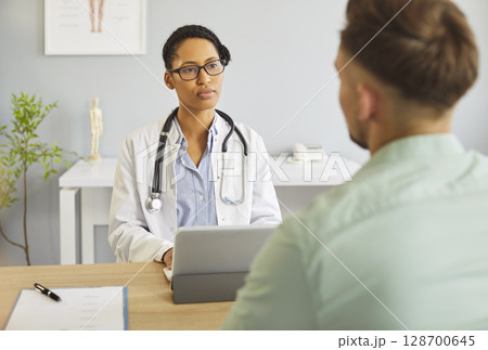 Young man patient talking to female doctor in her office in clinic during medical examination. Young man patient talking to female doctor in her office in clinic during medical examination. 128700645