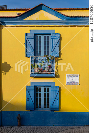 Colorful yellow building with blue shutters and a flower-filled balcony in a sunny village 128700656