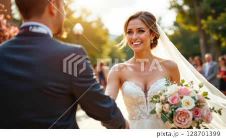 Joyful bride in beautiful wedding dress smiles at her groom, holding bouquet of roses in romantic outdoor setting 128701399