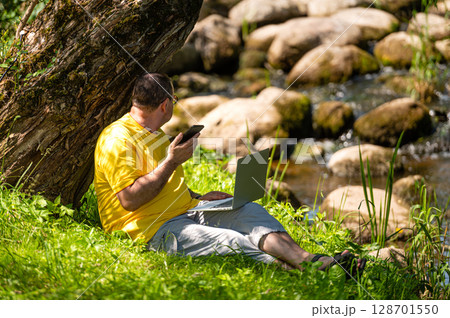 man with laptop and smartphone working outside in the meadow by the river, outdoor office concept 128701550