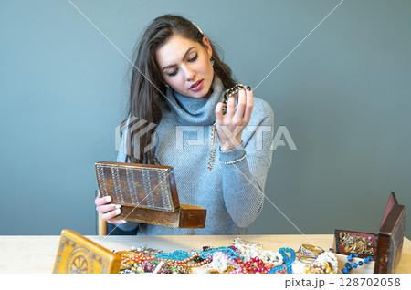 young brunette sitting at the table with lots of jewelry, choosing and trying different jewelry young brunette sitting at the table with lots of jewelry, choosing and trying different jewelry 128702058