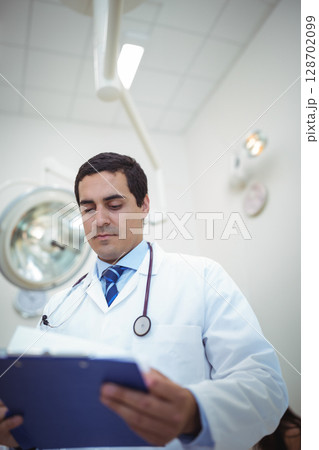 Reviewing blue clipboard male doctor wearing stethoscope in exam room, with surgical lamp and clock Reviewing blue clipboard male doctor wearing stethoscope in exam room, with surgical lamp and clock 128702099