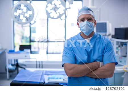 Standing male surgeon crossing arms in OR with surgical lights and blue drape, copy space Standing male surgeon crossing arms in OR with surgical lights and blue drape, copy space 128702101
