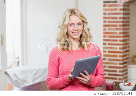 Smiling woman holding tablet in bright home kitchen, with ceramic mug and bowl of fruit 128702141