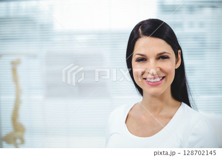Smiling woman standing indoors at medical clinic, with spine model and window blinds, copy space 128702145