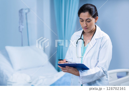 Asian female doctor reviewing notes on blue clipboard with stethoscope at hospital bed, copy space Asian female doctor reviewing notes on blue clipboard with stethoscope at hospital bed, copy space 128702161