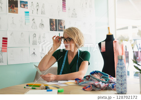 Adjusting senior woman reviewing paper sketches in fashion design studio, with colored pencils 128702172
