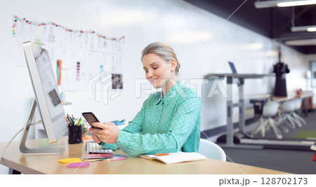 woman scrolling smartphone at desk in open office, with computer monitor and swatches woman scrolling smartphone at desk in open office, with computer monitor and swatches 128702173
