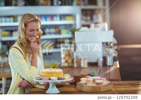 Admiring woman standing at bakery counter touching lips, with frosted cake and macarons, copy space 128702208