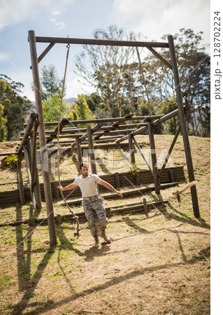 Climbing man navigating obstacle frame on wooded hillside, with hanging rope and log barriers Climbing man navigating obstacle frame on wooded hillside, with hanging rope and log barriers 128702224