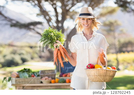Smiling woman holding fresh carrots and basket with peppers and tomatoes at farm stand 128702245