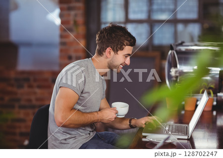 Man typing on silver laptop at industrial-style coffee shop counter, holding coffee mug 128702247