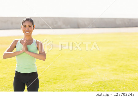 Smiling mid-adult Asian woman holding prayer gesture outdoors on lawn, with yoga focus, copy space 128702248