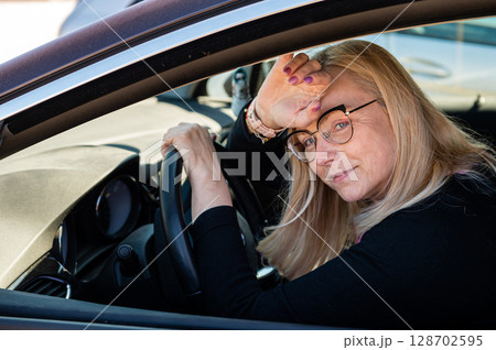 thoughtful middle-aged blonde in glasses sits behind the wheel of a car thoughtful middle-aged blonde in glasses sits behind the wheel of a car 128702595