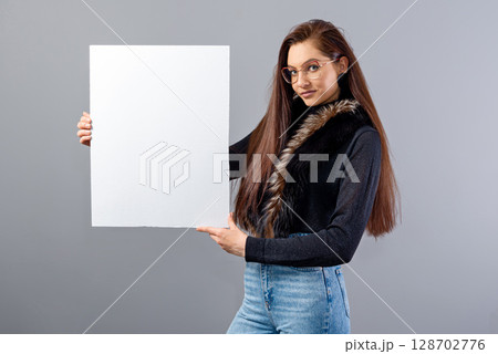 portrait of young teenager woman showing blank signboard with copy space, isolated on gray 128702776