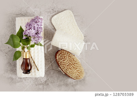Spa treatment flat lay with bottle of organic oil essence collagen. Dropper bottle with natural serum on a white towel with lilac flowers, massage brush and loofahs on grey background. Copy space Spa treatment flat lay with bottle of organic oil essence collagen. Dropper bottle with natural serum on a white towel with lilac flowers, massage brush and loofahs on grey background. Copy space 128703389