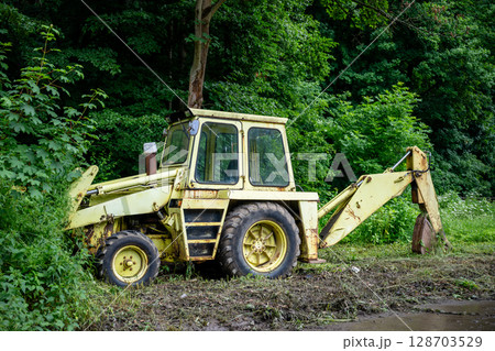 Old yellow backhoe loader parked at forest edge with overgrown vegetation Old yellow backhoe loader parked at forest edge with overgrown vegetation 128703529