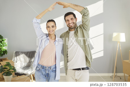 Happy young couple forming roof shape with hands over heads, celebrating purchase of new apartment. 128703535