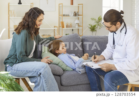 Male doctor writing prescription during home visit while child resting on sofa with mother nearby. 128703554