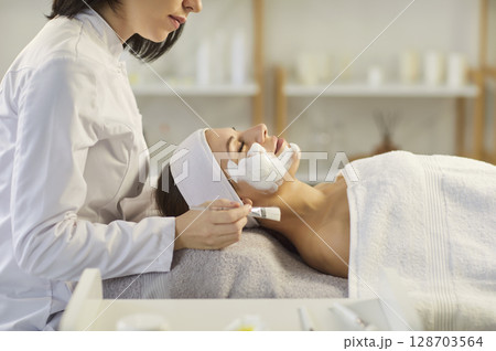 Professional cosmetologist applying moisturizing mask to young woman lying on couch in spa salon. 128703564