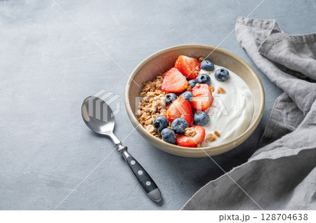 Natural yogurt with granola, blueberry and strawberry in a bowl on a blue background with spoon  128704638